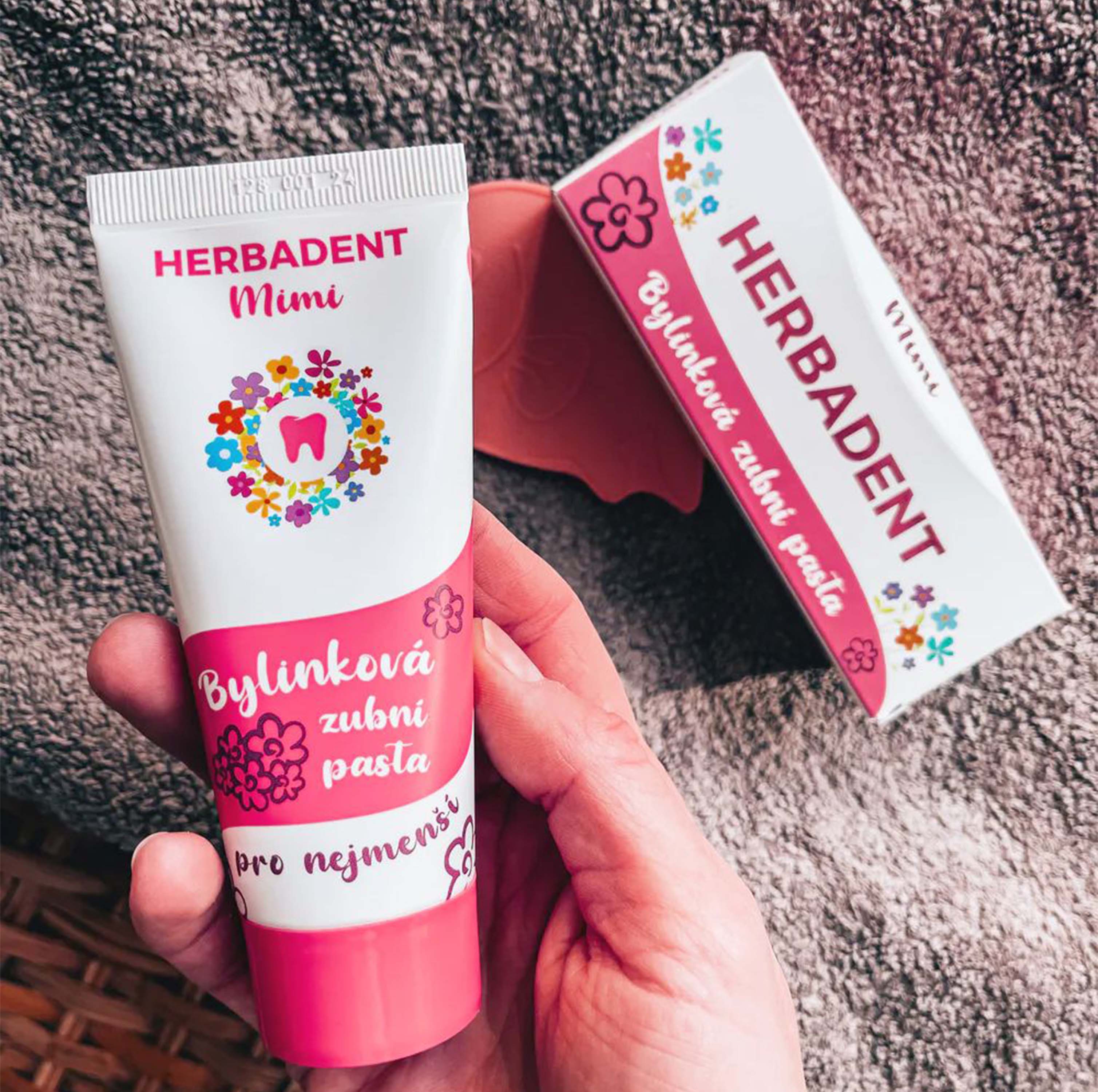 Hand holding a tube of Herbadent Mimi children's toothpaste with packaging in the background.