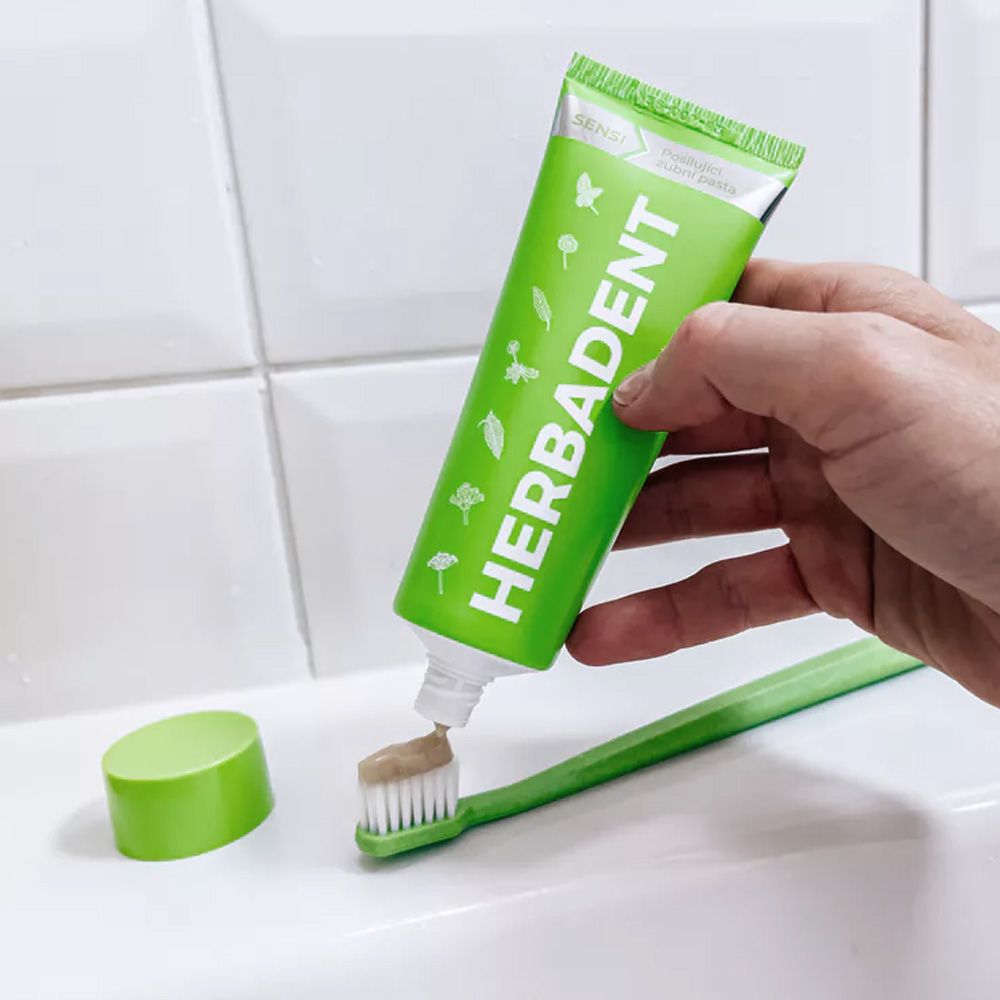 Hand holding a tube of Herbadent toothpaste with a toothbrush on a white surface.