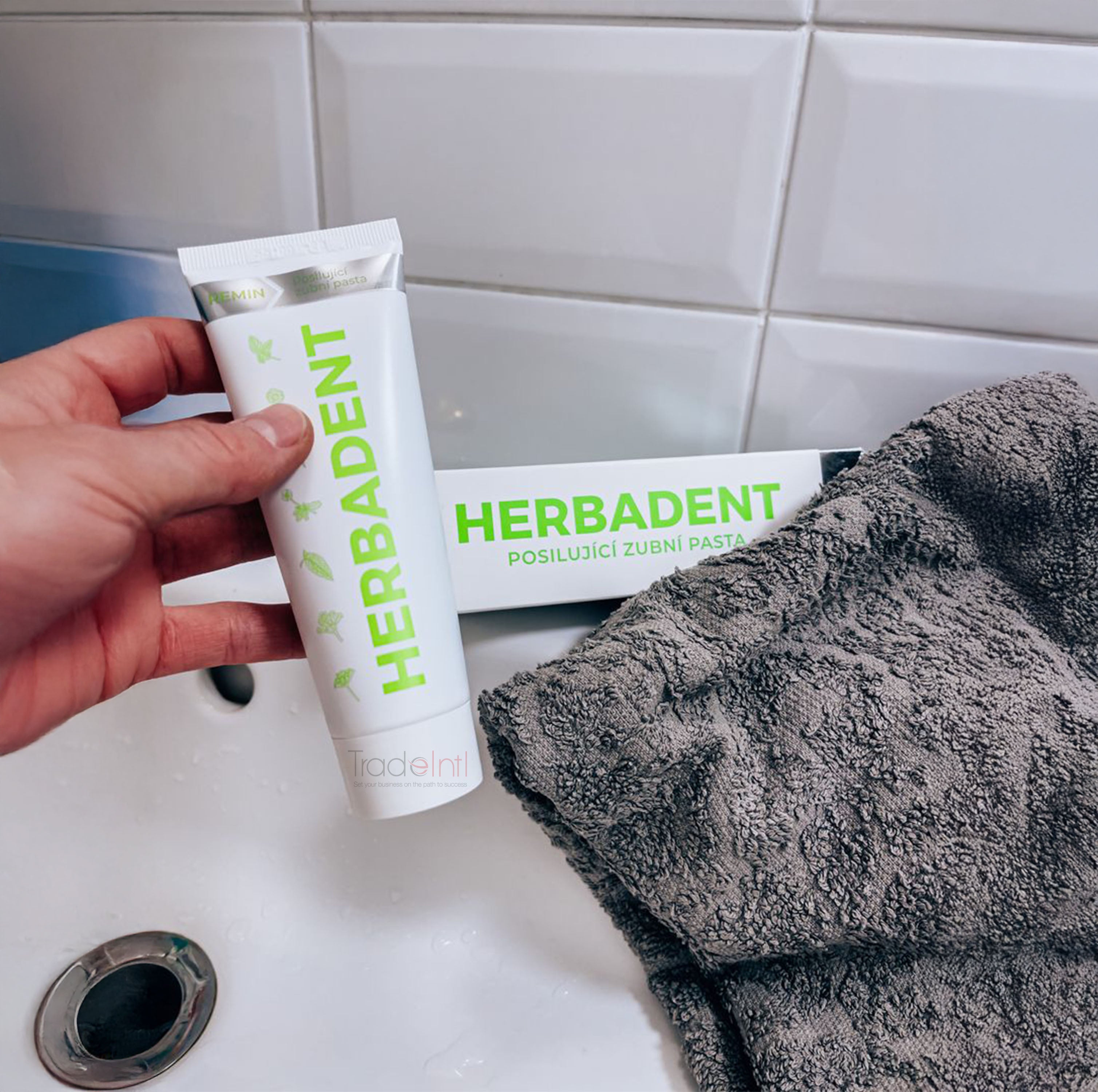 Hand holding a tube of Herbadent toothpaste with a towel on a sink.
